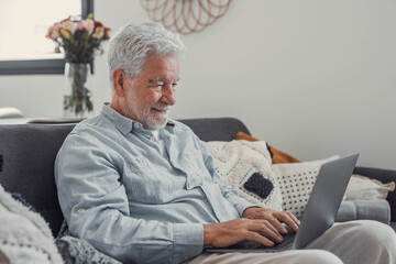 Elderly man sits on a sofa using his computer to browse and shop online. He enjoys a productive...