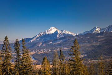 Snow-capped mountain range under a bright blue sky, towering over a valley of dense forest and small settlement. Zakopane town at the foot of the Tatra Mountains in Poland, Europe.