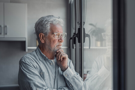 Elderly man looks out the window with concern. He appears thoughtful and worried about his life and surroundings while standing at home.