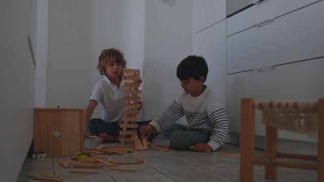 Two boys sitting in kitchen corner constructing tall wooden block tower with focused teamwork as scattered pieces surround them creating engaging playful childhood moment of patience and creativity