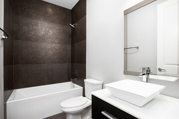 A modern bathroom with a vessel sink on a white countertop and wood cabinet and a dark brown floral tiled shower and bathtub.