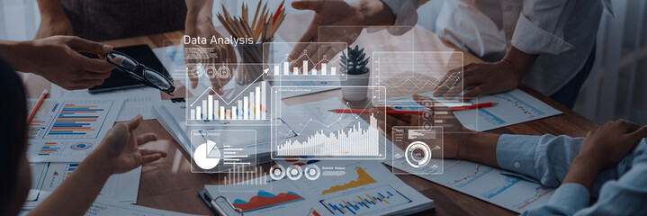 Business professionals engage in collaborative data analysis, discussing charts and graphs on a table filled with documents, tablets, and digital visuals while planning strategies. Trope