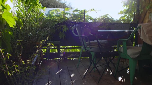 Balcony terrace with metal chairs and table surrounded by lush foliage, wooden deck partially shaded by leaves, tranquil outdoor rest space evoking peaceful gentle slow living atmosphere