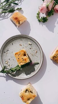 golden herb focaccia bread pieces scattered on and around a ceramic plate with crumbs, greenery, and soft floral accents on a white surface.