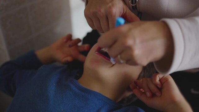 Mother brushing young boy&rsquo;s teeth while holding his jaw steady with close view of toothbrush motion foam and parental hand positions inside tiled bathroom environment