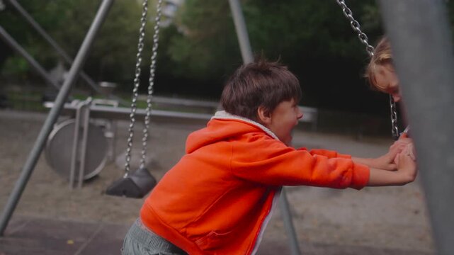 Older child pushing toddler seated in bucket swing at playground with visible chains metal frame and climbing structure in background during forward swing motion