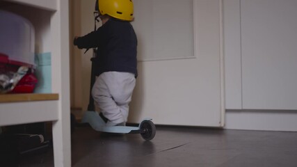 Toddler pushing scooter indoors wearing helmet moving with early balance confidence exploring home space with playful determination and gentle during childhood moment of fun