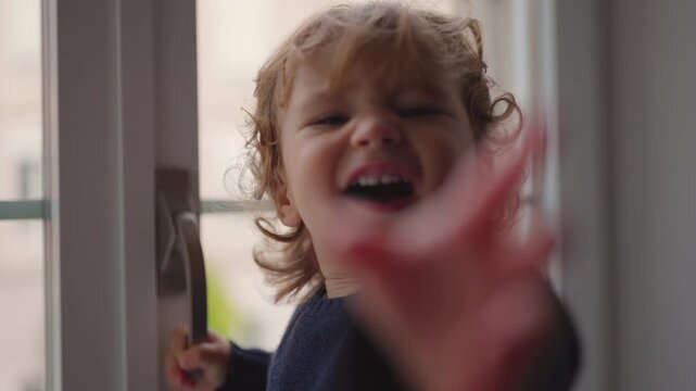 Toddler raising hand toward camera as if saying stop while standing by window showing strong expression playful assertiveness and emotional reaction