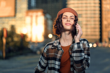 Fototapeta premium woman phone urban golden hour candid lifestyle beanie portrait: Young woman smiling on phone in city street at golden hour glow, casual fashion and curly hair, authenticity, mindful living and