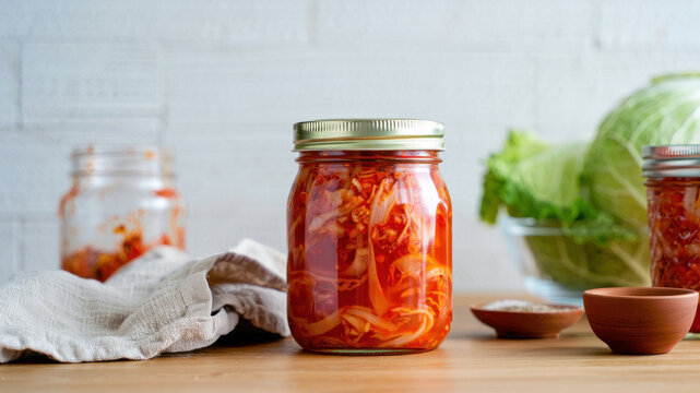 Homemade kimchi in glass jar on wooden table, spicy fermented cabbage with green leaves background, traditional Korean dish, probiotic-rich culinary tradition, healthy plant-based fermentation