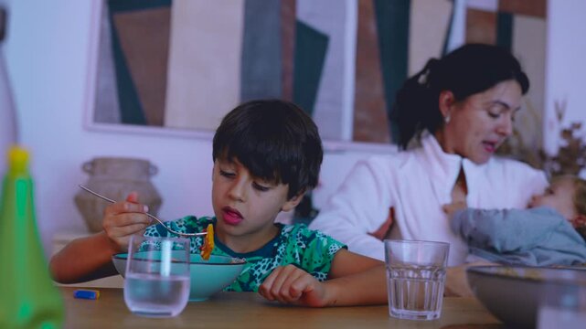 Mother holding toddler in her arms while older child eats pasta at table showing simultaneous family actions during everyday mealtime at home