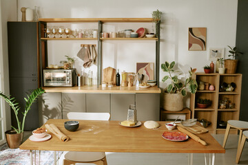 Wooden kitchen table holding rolling pin, dough, grated cheese, sliced sausage, and bowl of beans, kitchen shelves and potted plants visible in background