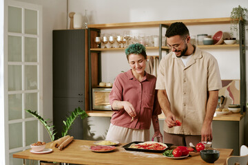 Young adult Caucasian woman and man preparing homemade pizza together in modern kitchen, smiling and interacting while arranging toppings on dough, fresh vegetables on table