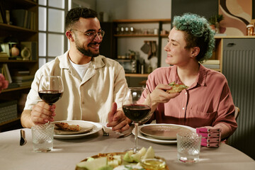 Young adult Caucasian woman with short curly hair and young adult man sitting at dining table eating together, holding wine glasses, smiling and making eye contact