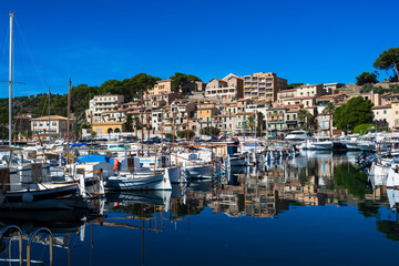 Coastal marina with moored boats, waterfront buildings and reflections on calm water on a sunny day.