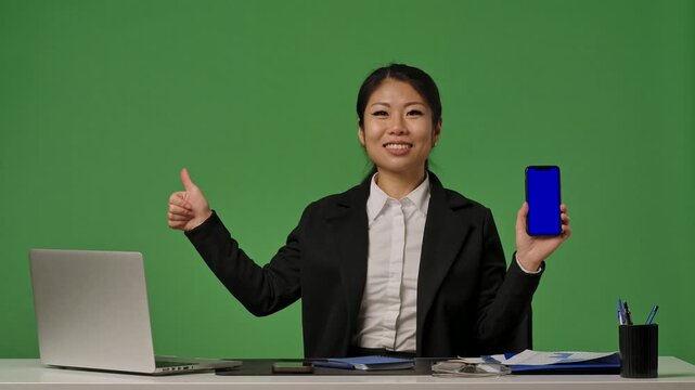 A young Korean businesswoman in a black suit holds a phone with a blue screen and makes a thumbs up gesture
