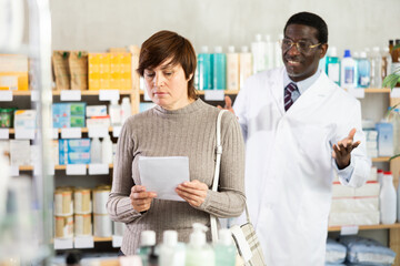 Obraz premium In sales area of drugstore, woman with prescription looks at display cases, trying to find multivitamins for adults in open display. African male pharmacist informs about possibility of replacing