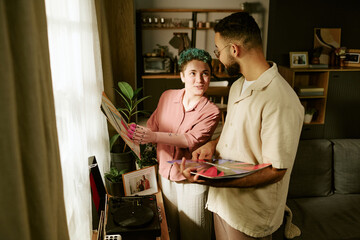 Caucasian young adult woman with prosthetic arm selecting vinyl record while smiling at man holding records, both standing near record player in living room