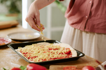 Caucasian young adult woman sprinkling shredded cheese over uncooked pizza dough on baking tray, preparing homemade pizza in kitchen, visible hand and partial torso