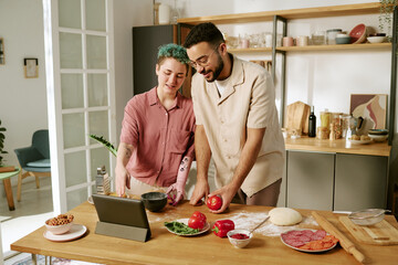 Young adult Caucasian woman and young adult Middle Eastern man preparing fresh ingredients together in kitchen, slicing vegetables and using digital tablet for recipe guidance