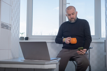 Man sits by window with laptop and phone while enjoying cup of orange drink on a cool morning in a bright kitchen