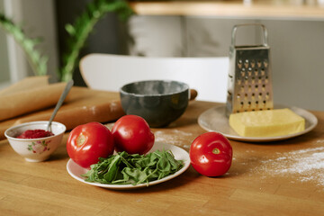 Fresh tomatoes and leafy greens resting on plate near cheese block and metal grater on wooden kitchen table, flour dusting surface, preparing ingredients for cooking meal