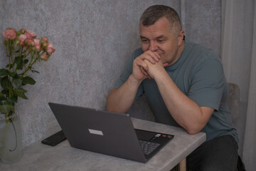 Man sitting at a table using a laptop with a vase of flowers nearby in a well-lit room during daytime