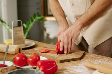 Unrecognizable man slicing fresh tomato on wooden cutting board in kitchen, hands covered with flour, surrounded by vegetables and kitchen utensils
