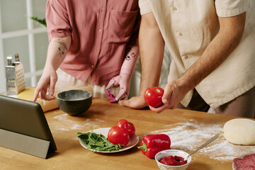 Caucasian young adult man and Caucasian young adult woman with prosthetic arm preparing food together in kitchen, following recipe on tablet, holding tomatoes and pointing at screen