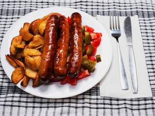 Fine dinner with long hot dog sausages, spicy potato wedges and simple country salad with marinated cucumbers, tomato on a white plate and black and white table cloth with fork and knife. Tasty meal