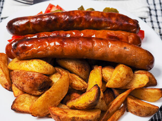 Fine dinner with long hot dog sausages, spicy potato wedges and simple country salad with marinated cucumbers, tomato on a white plate,