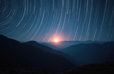 Star trails arc across deep blue night sky over silhouetted mountain ranges. Sun rises low on horizon, casting warm glow. Long exposure captures celestial movement, vastness, and natural beauty.