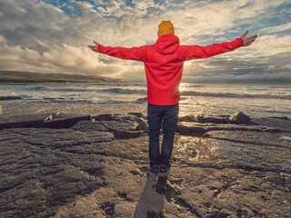 A man in a red jacket stands on a rock overlooking the ocean. The sky is cloudy and the water is...
