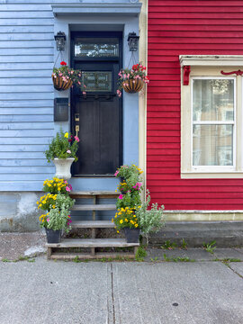 A blue house with a black door. There's a small window in the door, and over called a transom window. The adjoining house is vibrant red with white trim. The steps to the door have colorful flowerpots