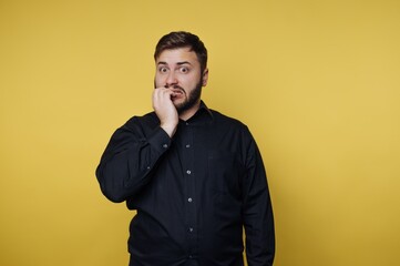 Fototapeta premium Man shows surprise and concern with hand on mouth in front of yellow background during daytime in indoor setting