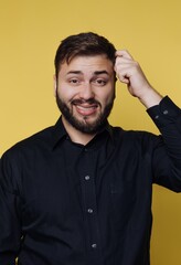 Obraz premium Man with beard and black shirt scratching head in front of yellow background while smiling during indoor setting
