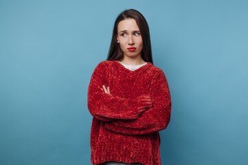 Young woman stands against blue wall with crossed arms, showing a thoughtful expression while...