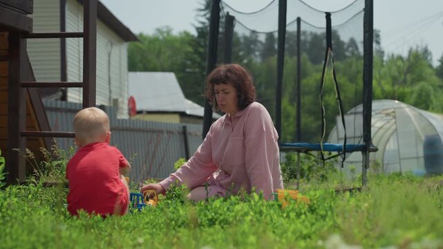 Mother And Child Play, Bright Day With Playful Bonding, Child And Mother Share Joyful Moments In Sunny Yard, Cheerful Woman Interacts Lovingly With Her Toddler Amidst Toys On Grassy Field