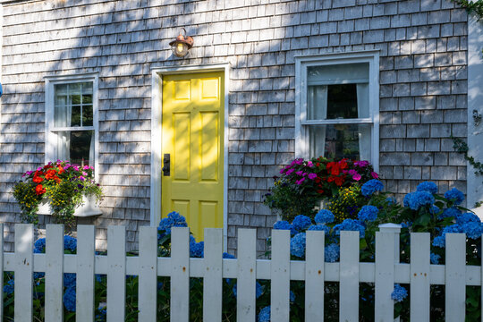 The exterior entrance to a small country cottage with grey cedar shakes, double hung windows, and a bright yellow solid panel door. There are colorful flowers, shrubs, and flower boxes in the yard.