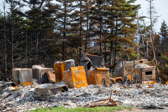 The charred remnants of a burnt residential building are the result of a wildfire. The trees and ground are scorched. There are ashes, a damaged car, and blackened property from the fire damage.