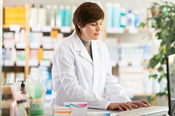 Focused middle-aged female pharmacist in white coat using desktop computer at pharmacy counter, checking medications or managing inventory