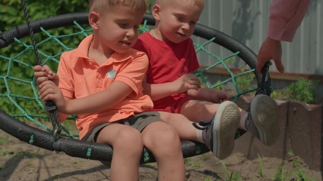 Children Enjoy Swinging Happily Outside, Youngsters Sit On Circular Swings In Sunlight, Happy Children Sit Peacefully On Round Swings While Adult Gently Adjusts And Pushes In Daylight