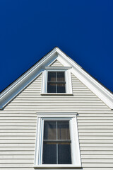 A peaked roof on a white wooden horizontal clapboard framed cottage. The historic building has exterior wood siding, two double hung glass windows, white trim boards with a dark blue sky background. 