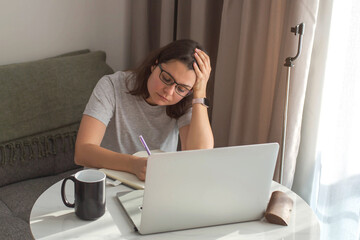 Tired woman working from home, sitting at a table with a laptop and writing in a notebook, holding her head in concentration while dealing with stress and deadlines in a cozy home workspace.