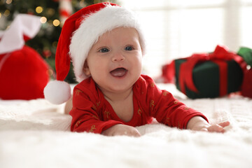 Cute baby in Santa hat on bed against blurred lights indoors. Christmas season