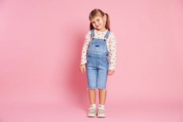 Cute little girl in jumpsuit standing on pink background