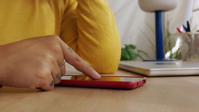 Close-up of a tattooed woman's hand using a red smartphone, scrolling through the screen while sitting at a wooden desk with a laptop and a lamp in the background in her modern home office