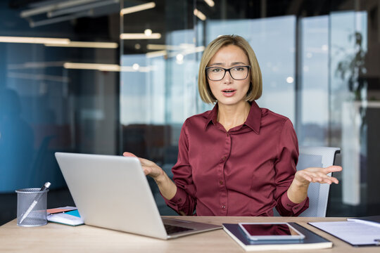 Businesswoman sitting at her desk with a laptop, shrugging with open hands, conveying confusion and frustration while dealing with a problem at work in a modern office