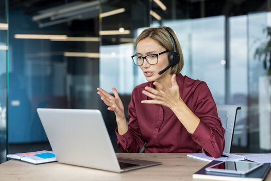 Businesswoman wearing a headset and glasses, actively participating in a video call on her laptop, gesturing with hands while communicating or troubleshooting a problem during a remote work session