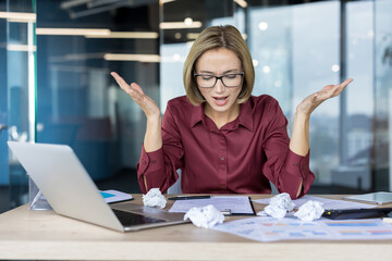 Young businesswoman feeling stressed and overwhelmed, sitting at her office desk with crumpled...